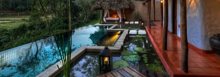 Rustic luxury villa veranda with red terracotta tiles leading to a turquoise infinity pool, lily pad pond with stepping stones, and a thatched daybed cabana at dusk